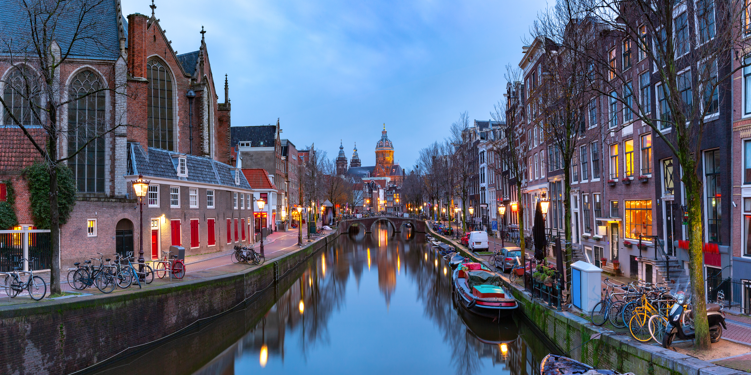 Amsterdam canal at dusk lined with gabled houses, bicycles and moored boats, a domed church in the distance